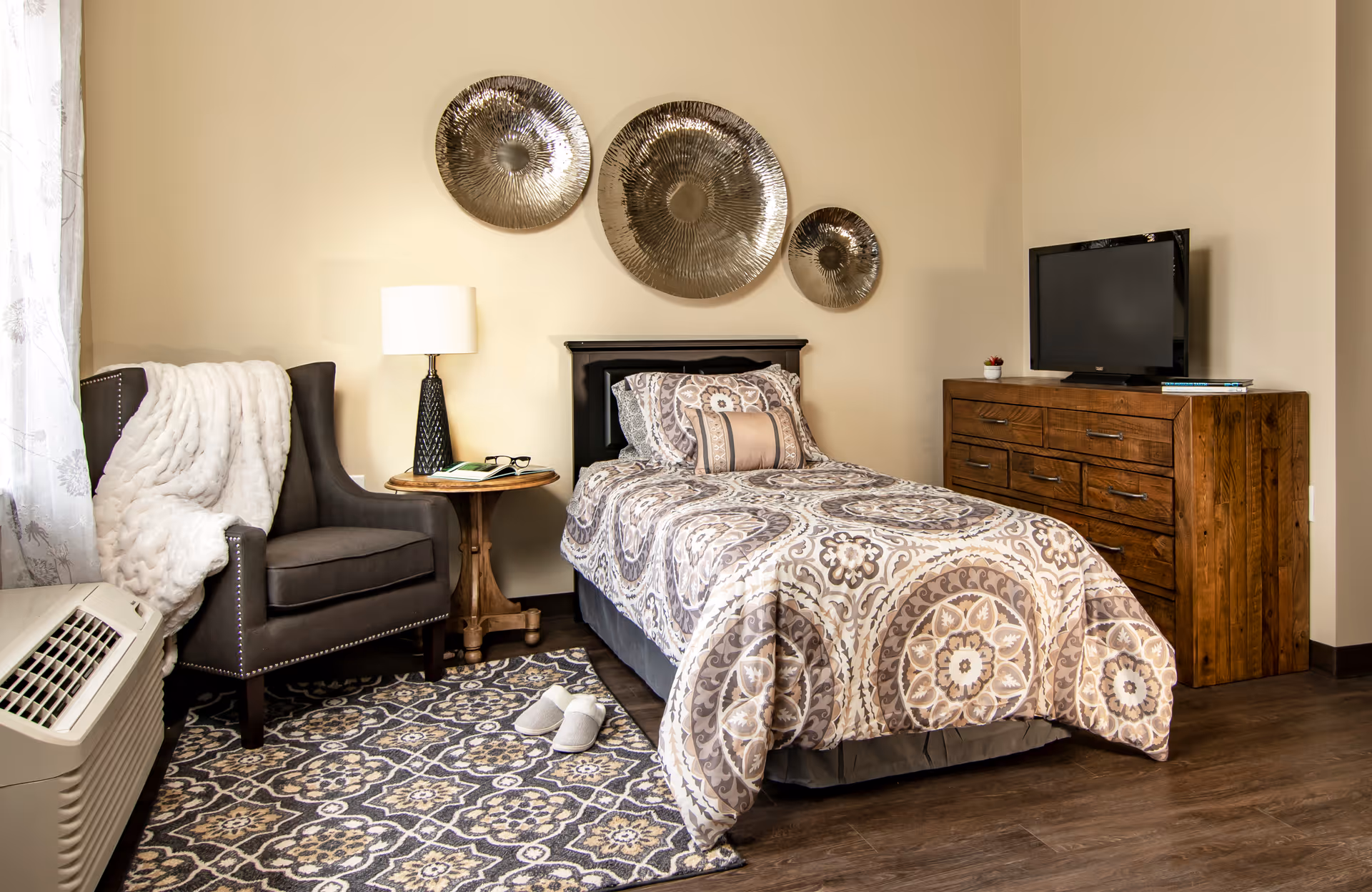 A cozy bedroom in a senior living community featuring a single bed with patterned bedding, a wooden dresser with a TV on top, a small round wooden side table with a lamp and glasses, a gray armchair with a white throw blanket, and a patterned rug on a wooden floor.