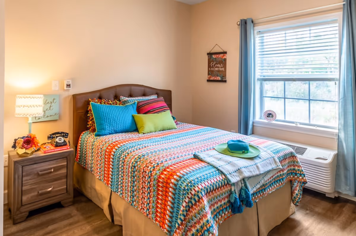 A cozy bedroom with a bed covered in a colorful striped blanket and multiple pillows in blue, green, and pink. Next to the bed is a wooden nightstand with a lamp, a vintage black rotary phone, and some decorative items. A window with blue curtains lets in natural light, and a wall hanging with floral design and the word 'Home' is visible on the wall.