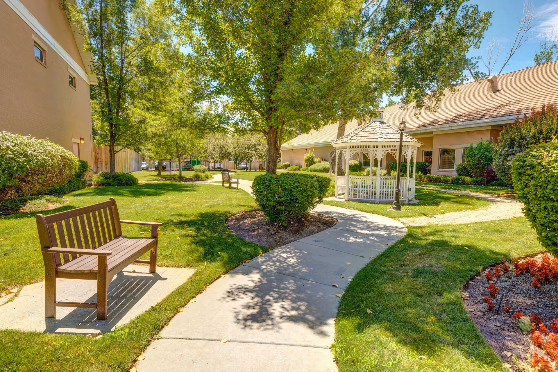 A sunny outdoor garden area with a paved walkway, wooden benches, green grass, trees, bushes, and a white gazebo near a beige building.