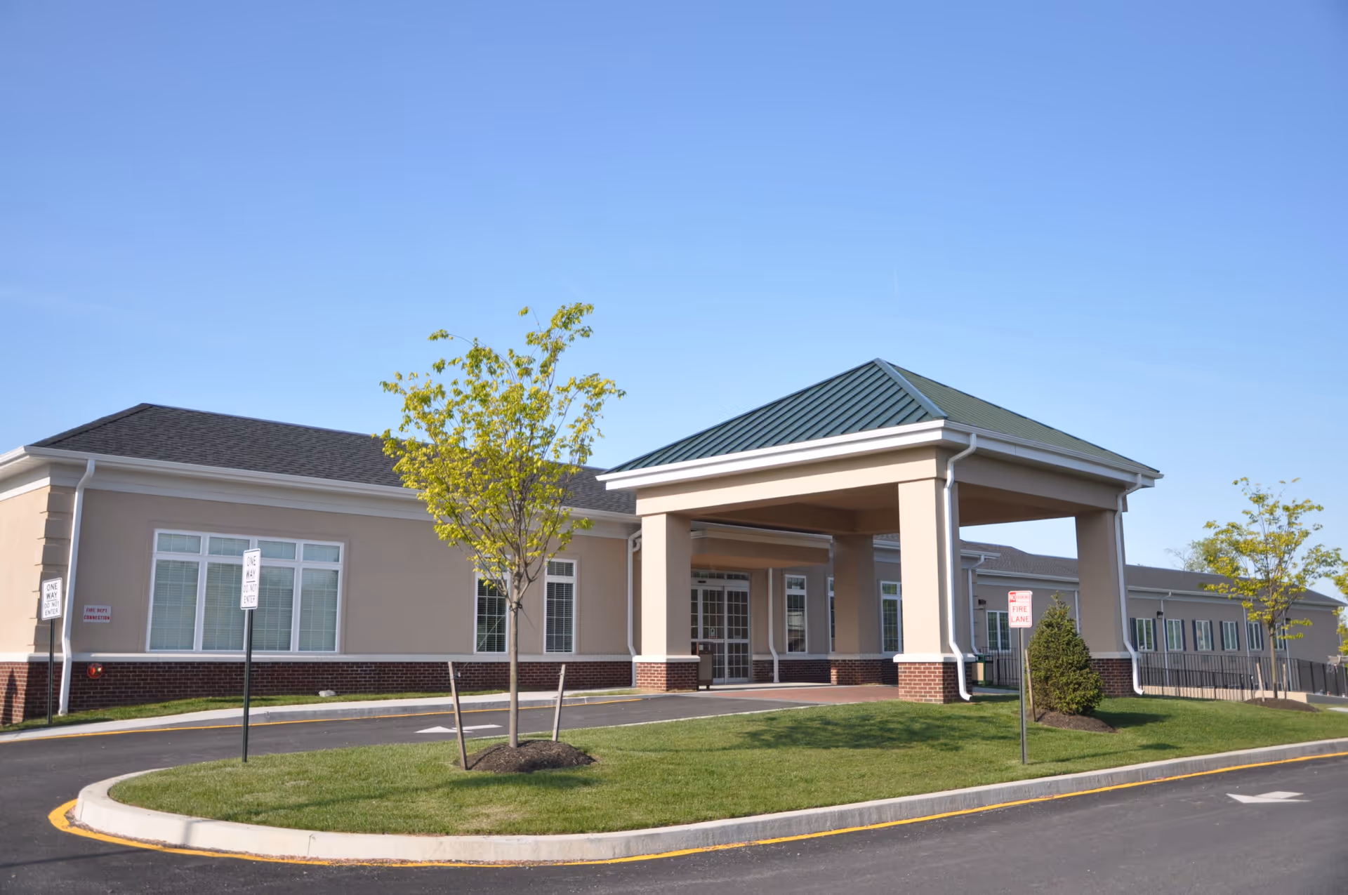 Exterior view of Cadia Rehabilitation Pike Creek building with a covered entrance, beige walls, brick accents, several windows, young trees, and a clear blue sky.