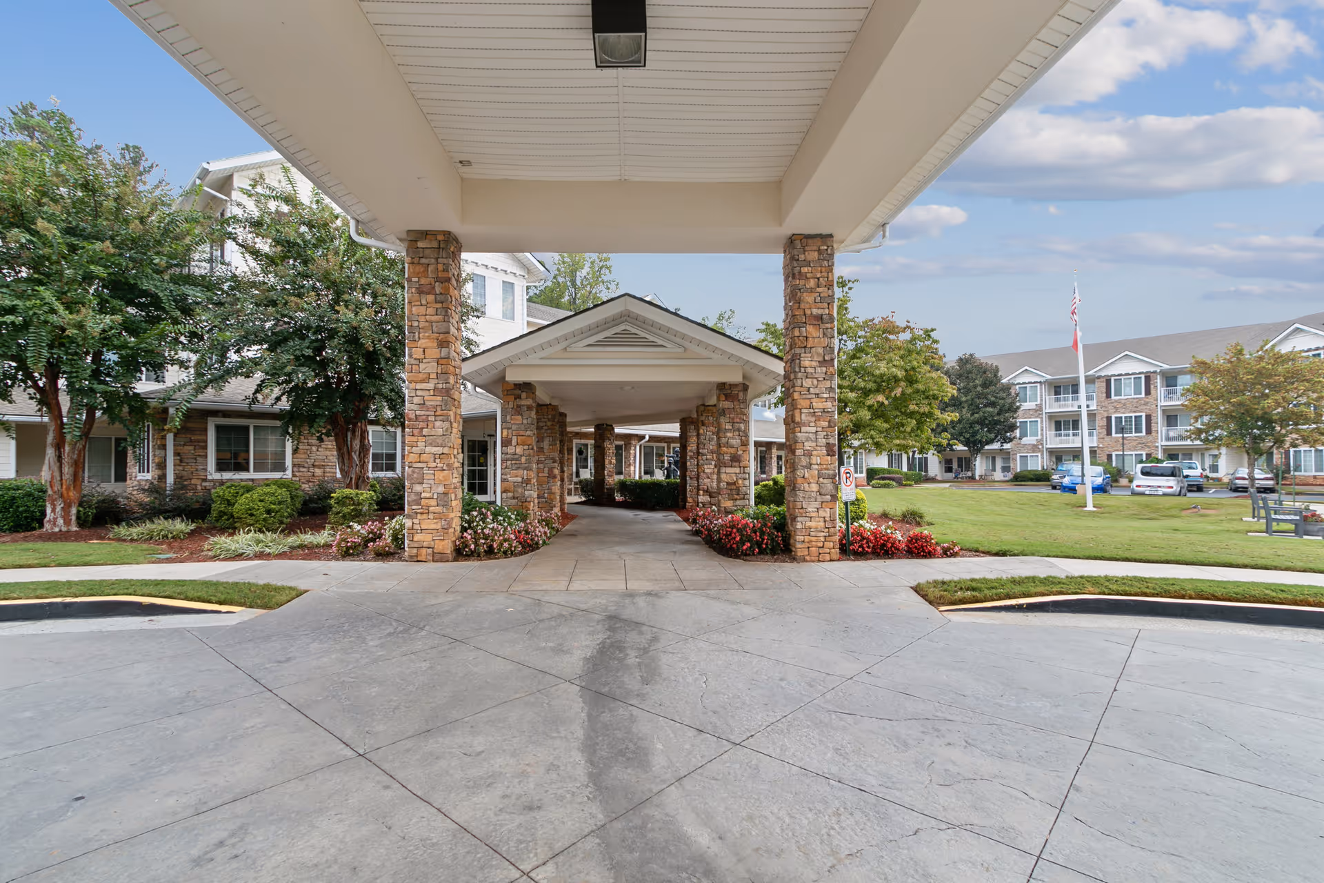 Covered porte-cochère with stone columns over a driveway leading to the entrance of a multi-story senior living building set in landscaped grounds.