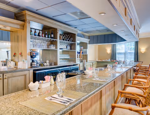 Interior view of a well-lit bar area with a long granite countertop, wooden bar stools with armrests, and shelves stocked with glasses and bottles. The setting includes place settings with napkins, utensils, and glasses, suggesting a dining or social area.