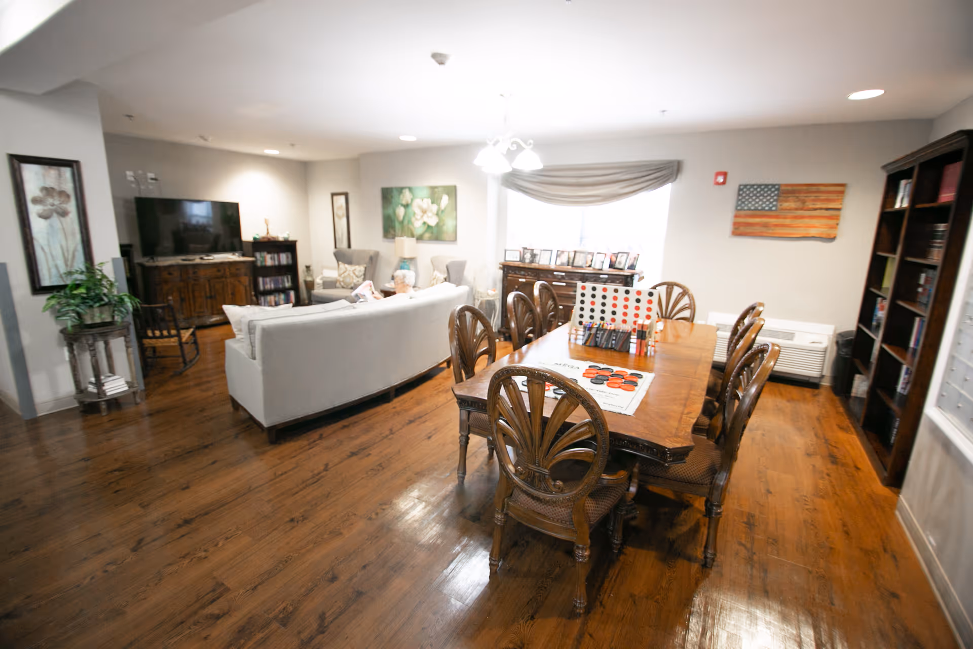 A cozy senior living common area with a wooden dining table and eight chairs in the foreground, featuring a large Connect Four game on the table. In the background, there is a seating area with a white sofa, two armchairs, a wooden TV stand with a flat-screen TV, and bookshelves. The room has wooden flooring, framed artwork on the walls, and a window with a curtain letting in natural light.