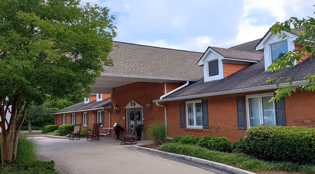 Front entrance of a single-story brick senior living building with a covered driveway, rocking chairs, and landscaped shrubs.