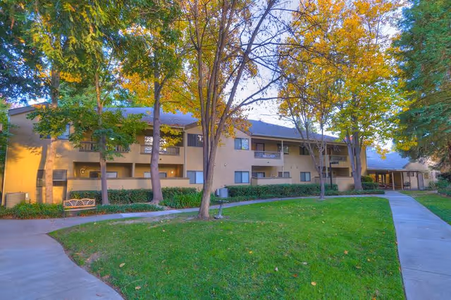 Exterior view of a two-story assisted living facility building surrounded by trees with autumn foliage. There is a well-maintained lawn and a curved concrete walkway leading to the entrance of the building.