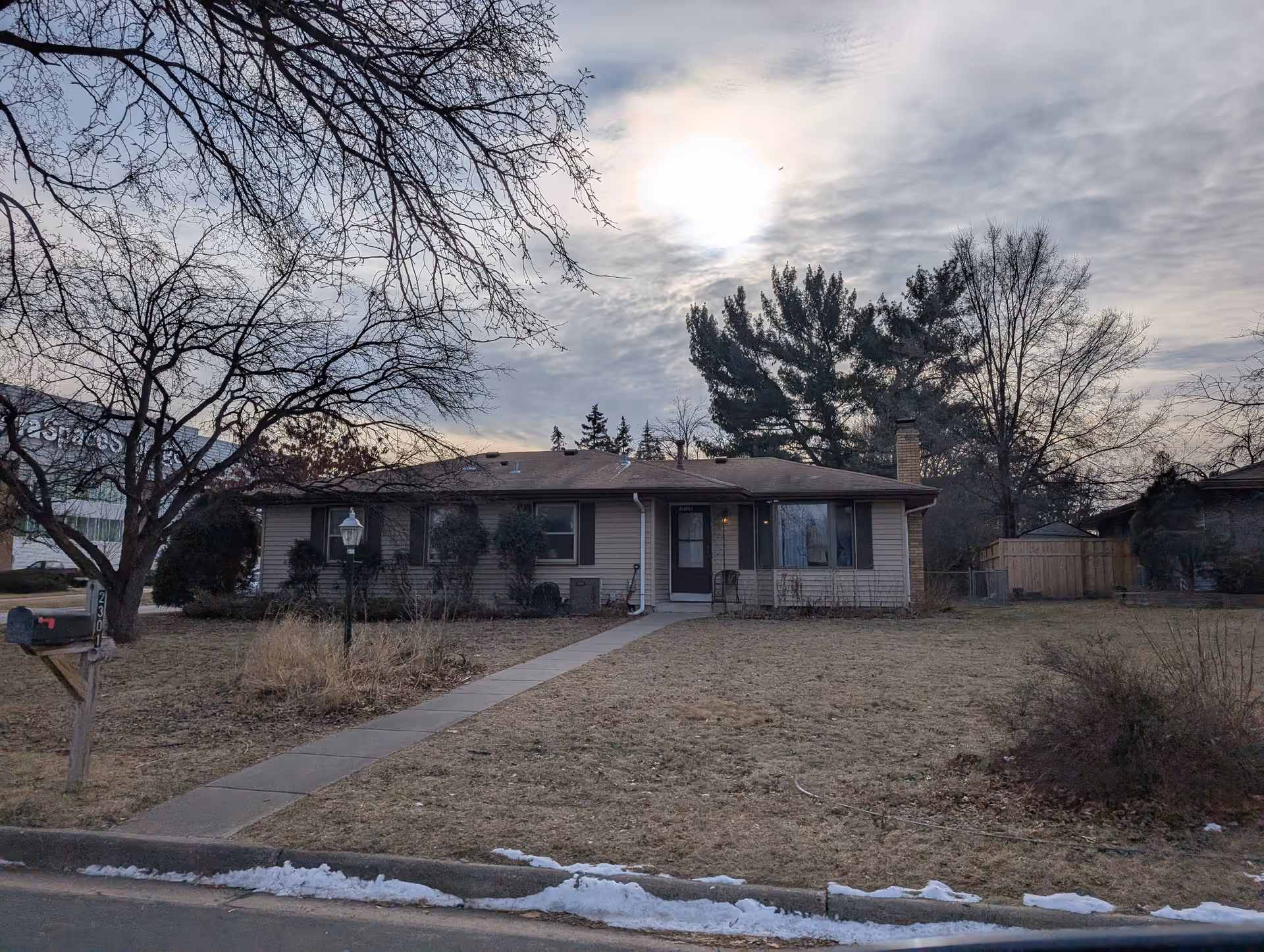 Single-story house with a brown roof and beige siding, a concrete walkway leading to the front door, leafless trees, a mailbox with the number 2301, and patches of snow on the ground under a cloudy sky with the sun partially visible.