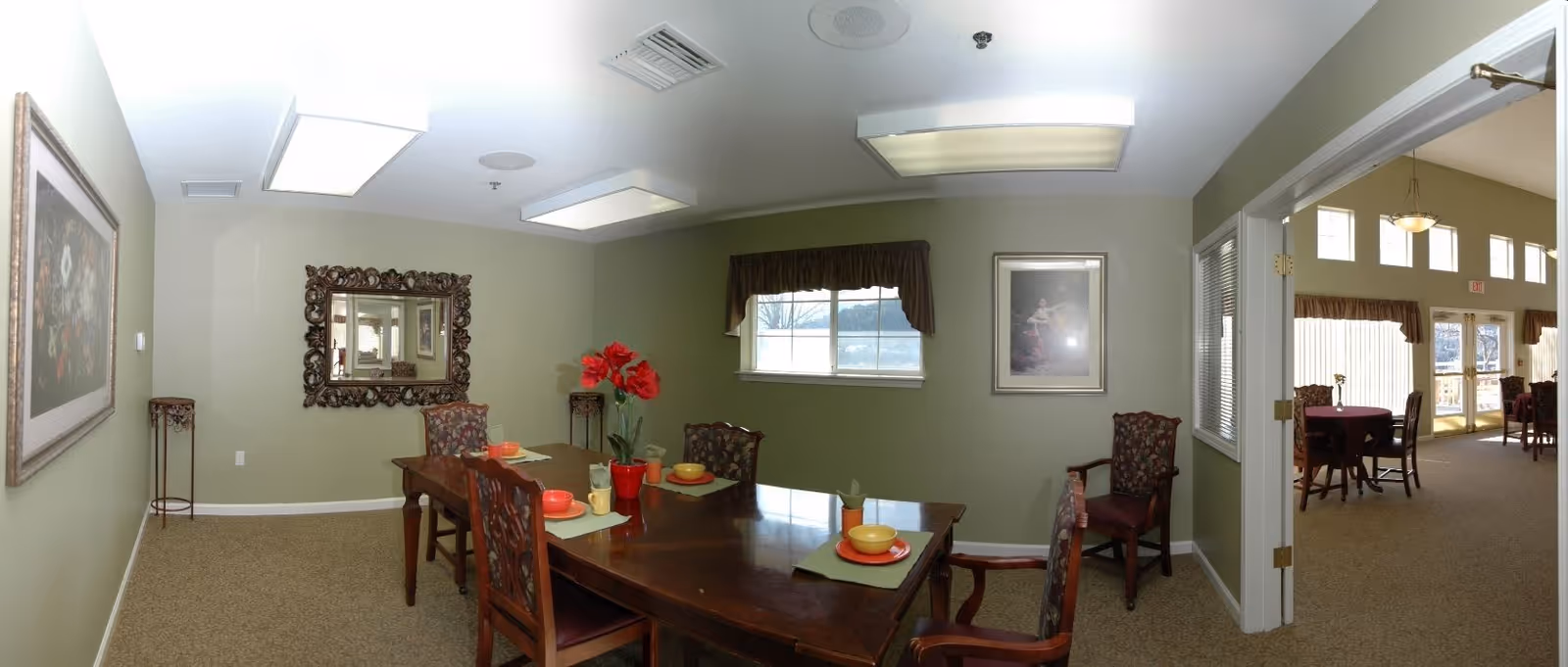 A dining room with a wooden table set for four with colorful bowls and cups, a vase with red flowers in the center. The walls are painted light green with framed artwork and a decorative mirror. There is a window with a brown valance and an adjoining room visible through an open doorway, showing more tables and chairs with large windows letting in natural light.