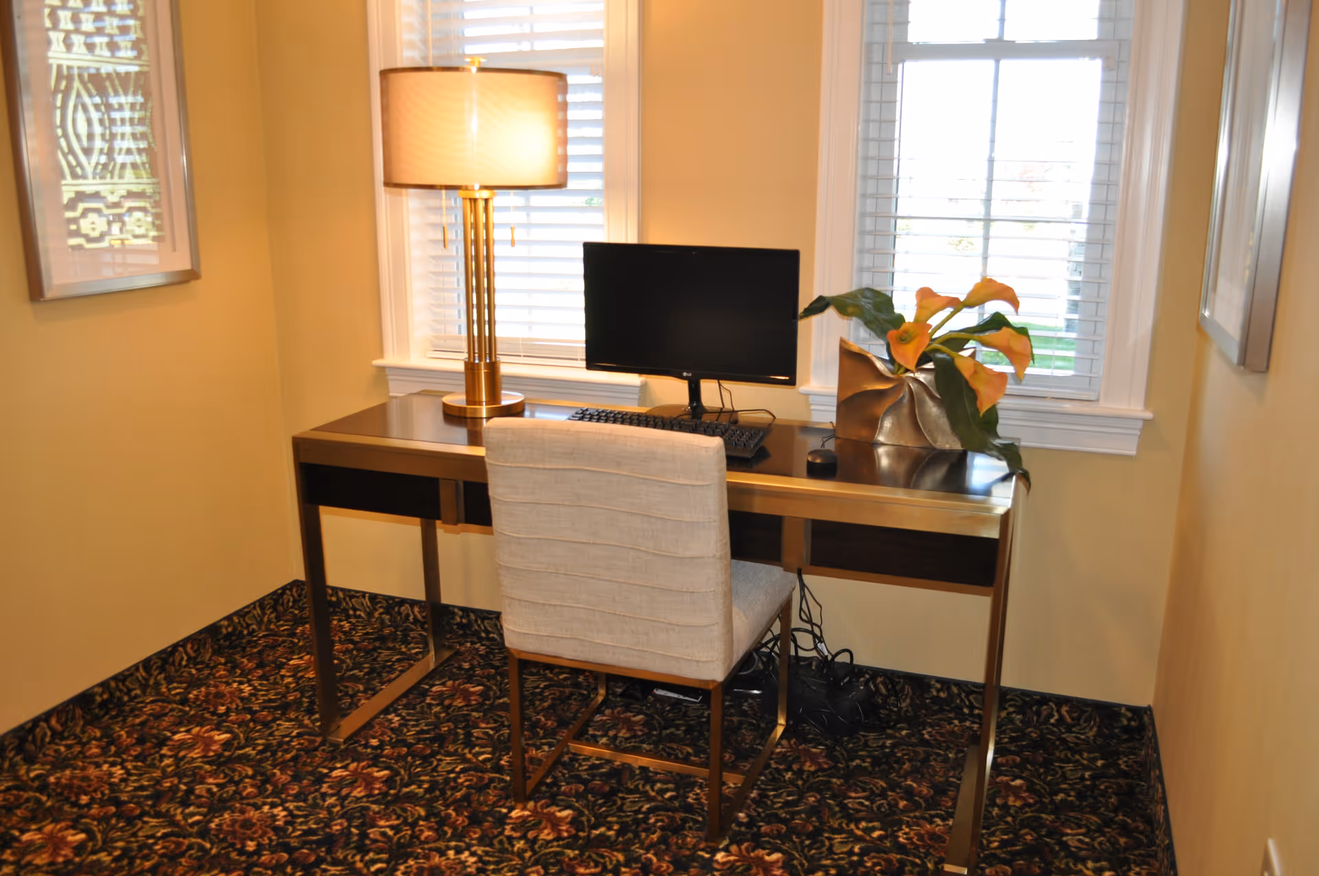 A small office nook with a modern desk and a beige upholstered chair. On the desk is a computer monitor, keyboard, mouse, a gold-colored table lamp with a beige shade, and a decorative vase with orange calla lilies. The area has two windows with white blinds and patterned carpet flooring.