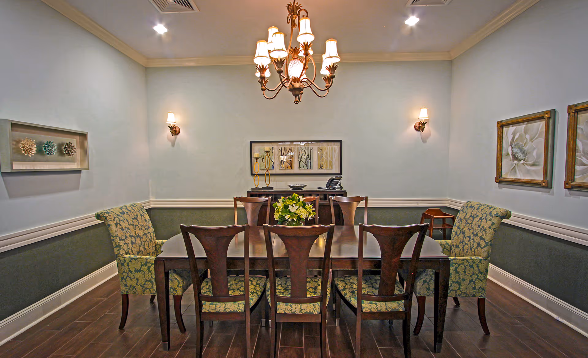 A formal dining room with a rectangular wooden table surrounded by six chairs with floral upholstery. The room has light blue walls with a darker green wainscoting, decorative wall art, a chandelier hanging from the ceiling, and two wall sconces providing warm lighting.