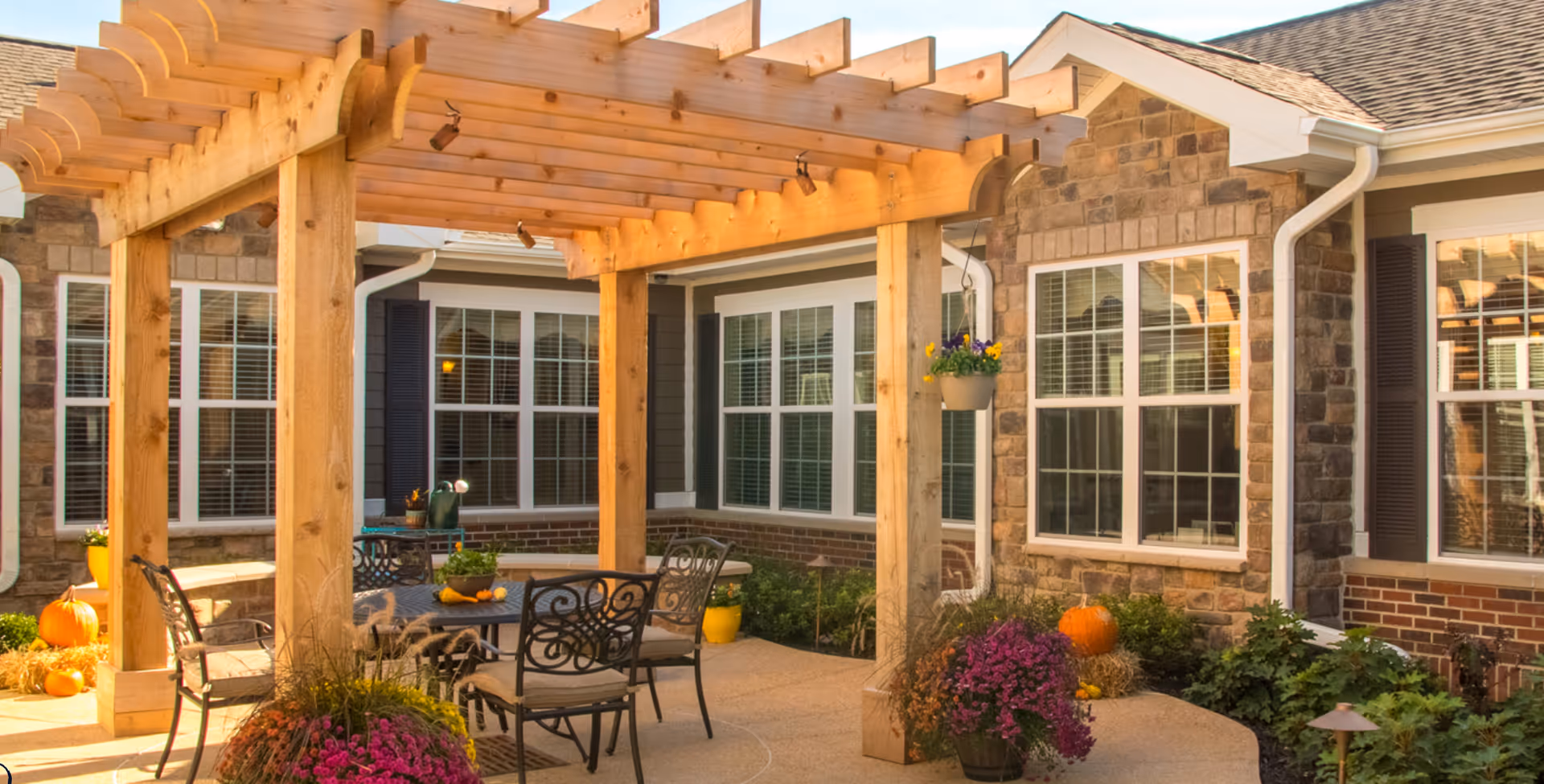 Outdoor courtyard patio with a wooden pergola, metal table and chairs, potted plants and pumpkins by the building's stone exterior and windows.