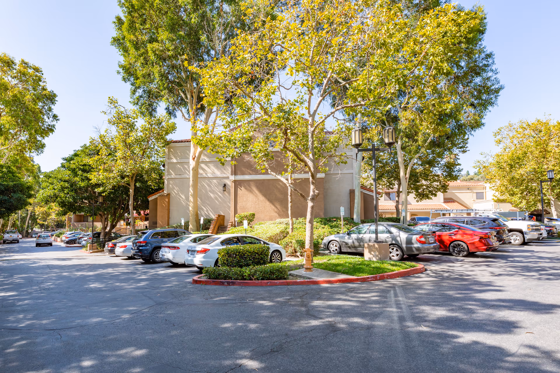 Parking lot with multiple parked cars surrounded by trees and greenery on a sunny day, with a beige building in the background.