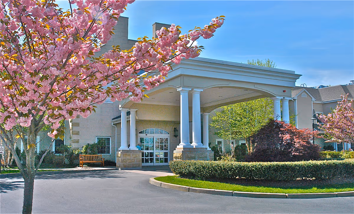 Front entrance of a beige senior living facility with a columned porte-cochere and blooming pink trees.