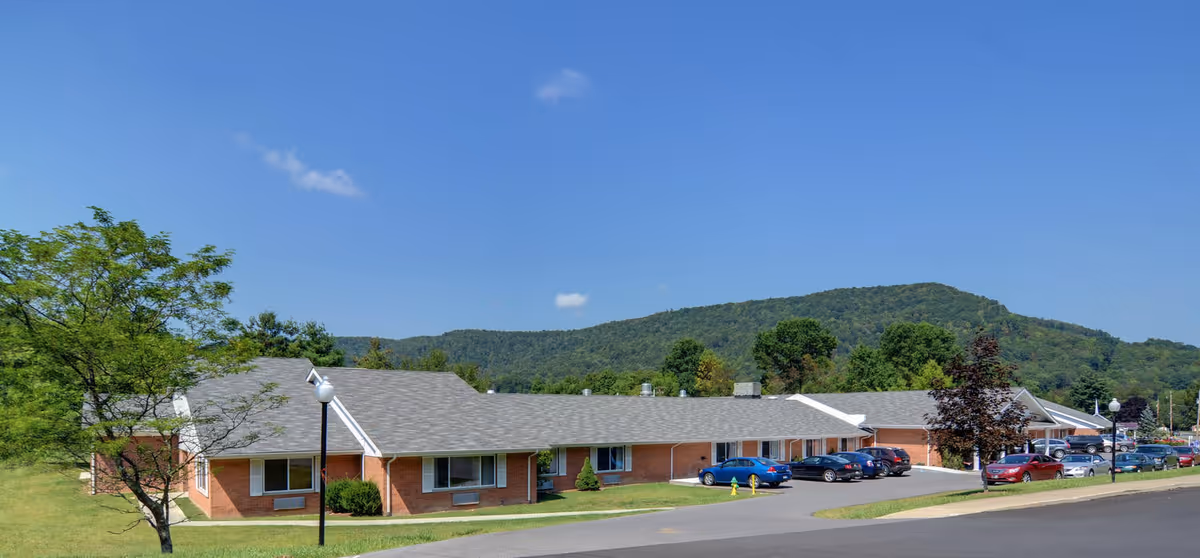 Single-story brick senior care building with a parking lot, lawn, trees and hills in the background under a clear blue sky.