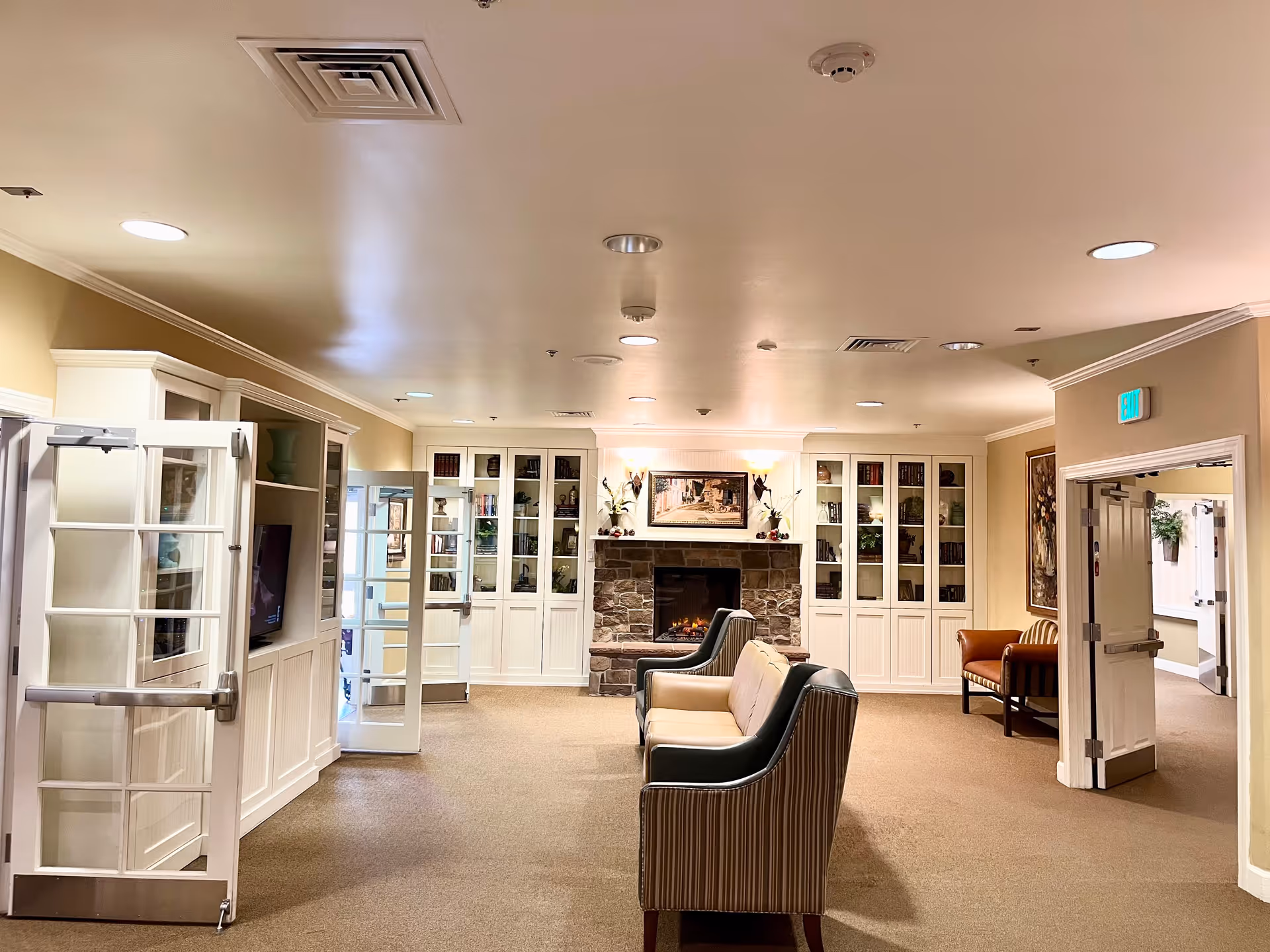 A cozy assisted living facility common area with a stone fireplace centered on the far wall, flanked by built-in white cabinets filled with books and decorative items. In front of the fireplace are two cushioned armchairs and a loveseat arranged for conversation. The room has beige walls and carpet, recessed ceiling lights, and glass double doors on the left side. There is an exit sign above a door on the right side leading to a hallway.