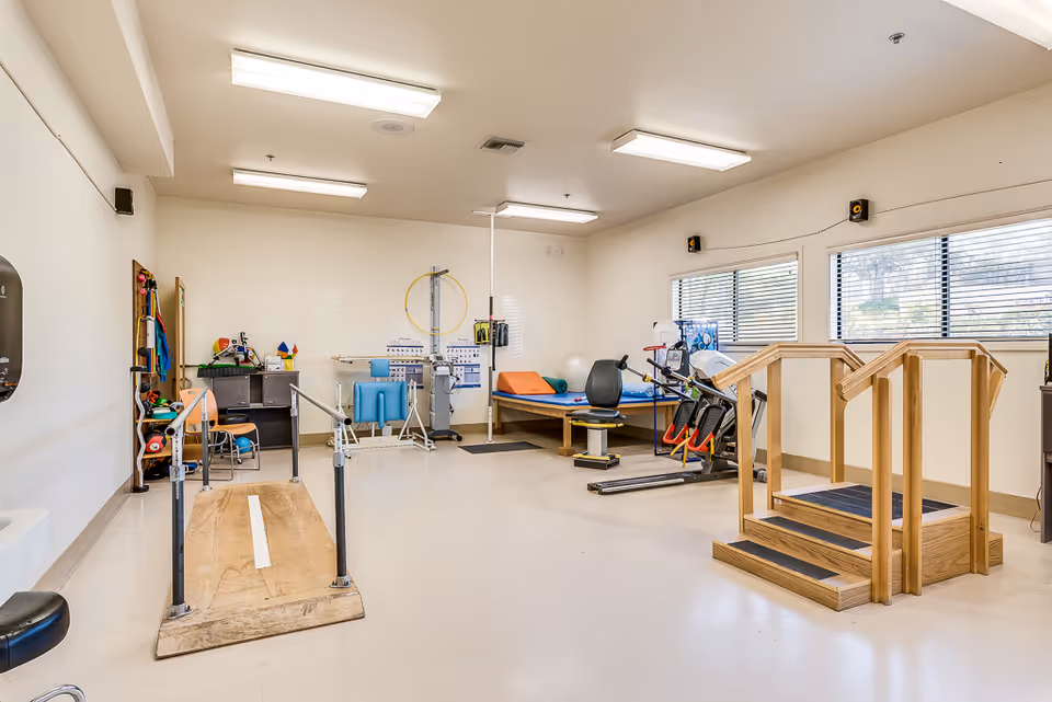Bright physical therapy room with parallel bars, wooden practice stairs, exercise equipment, treatment tables, and therapy supplies.
