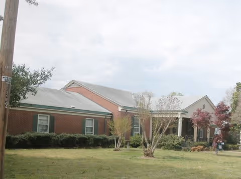 Exterior view of a single-story brick building with a gray roof, green window shutters, and a small covered porch. The building is surrounded by a grassy lawn with some bushes and small trees under a cloudy sky.