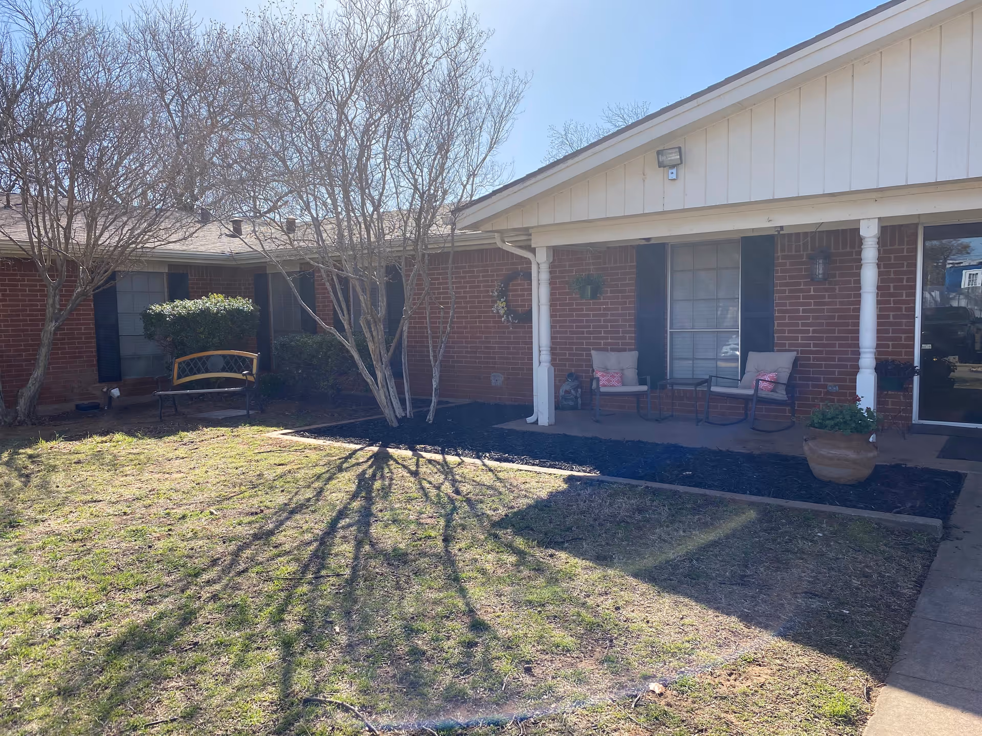 Outdoor view of a single-story brick building with a small porch area featuring two cushioned chairs and a small table. There are leafless trees and bushes in front of the building, along with a bench on the left side. The lawn is partially shaded with some grass and a paved walkway leading to the porch.