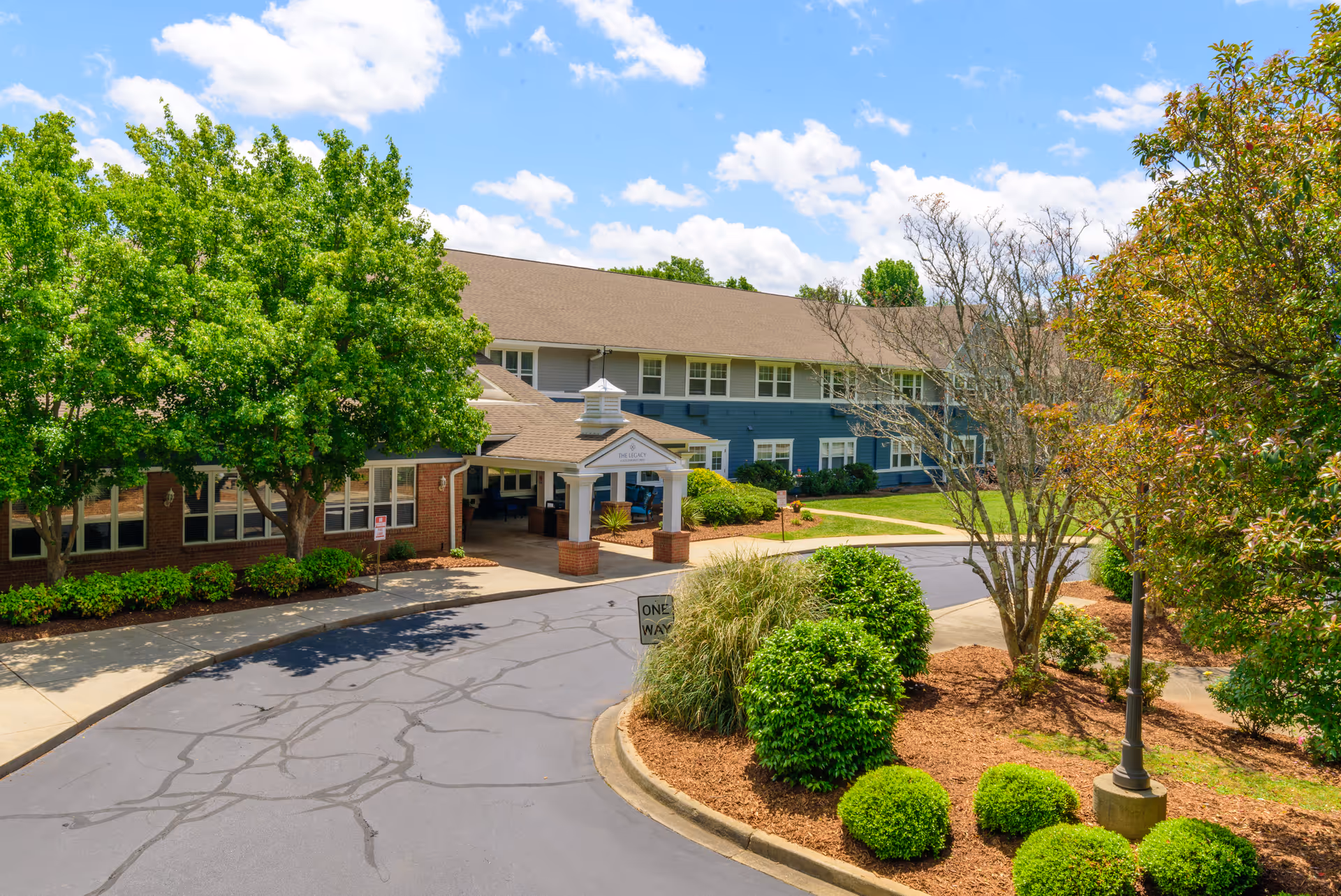 Exterior view of The Legacy at Southpointe Drive senior living facility showing a two-story building with a covered entrance, surrounded by green trees, shrubs, and a curved driveway under a partly cloudy blue sky.