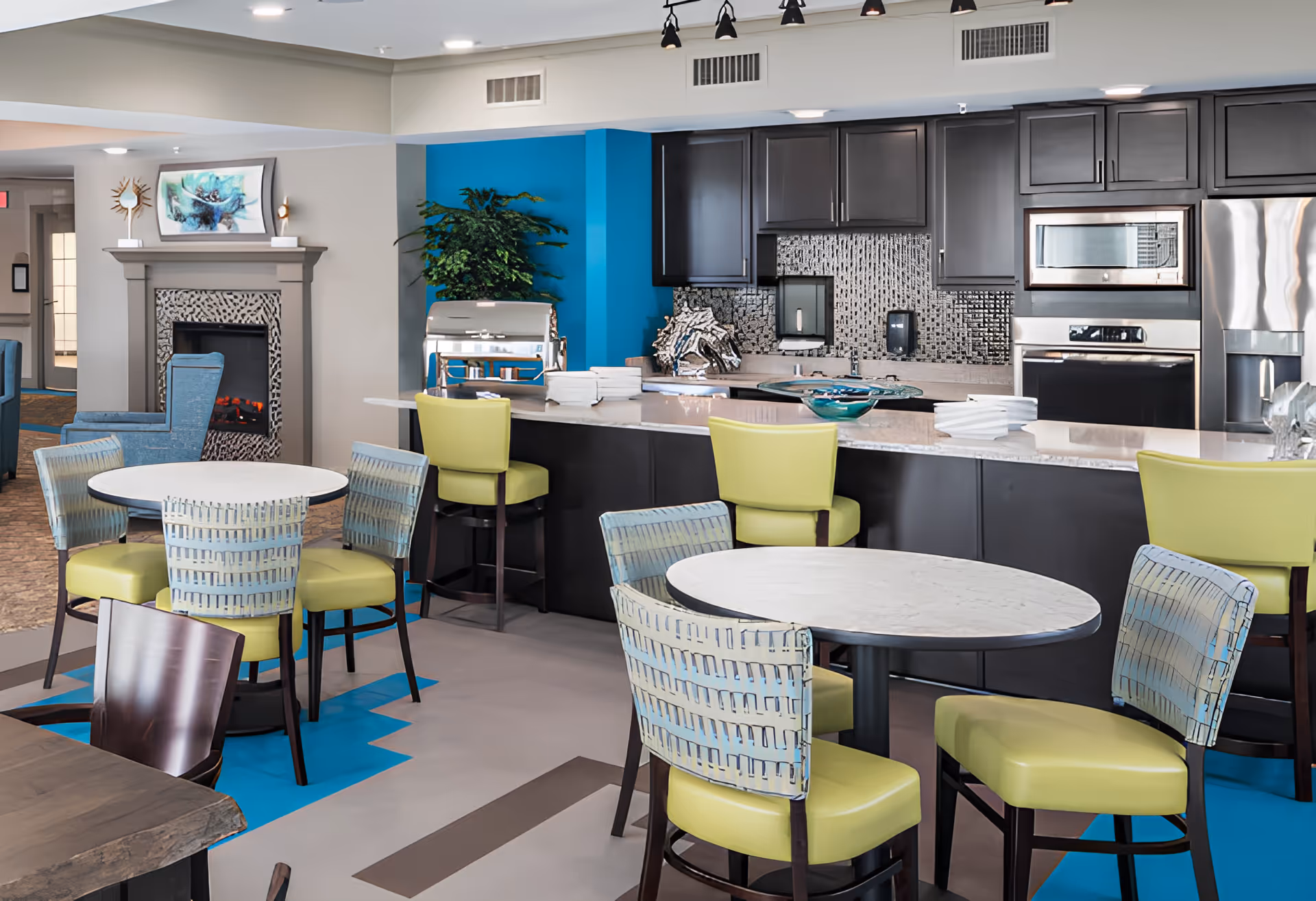 A modern dining area in a senior living facility featuring round tables with green and patterned chairs, a kitchen area with dark cabinets, stainless steel appliances, and a countertop with bar stools. There is a fireplace with a decorative mantle and artwork above it, and a blue accent wall with a plant near the kitchen.