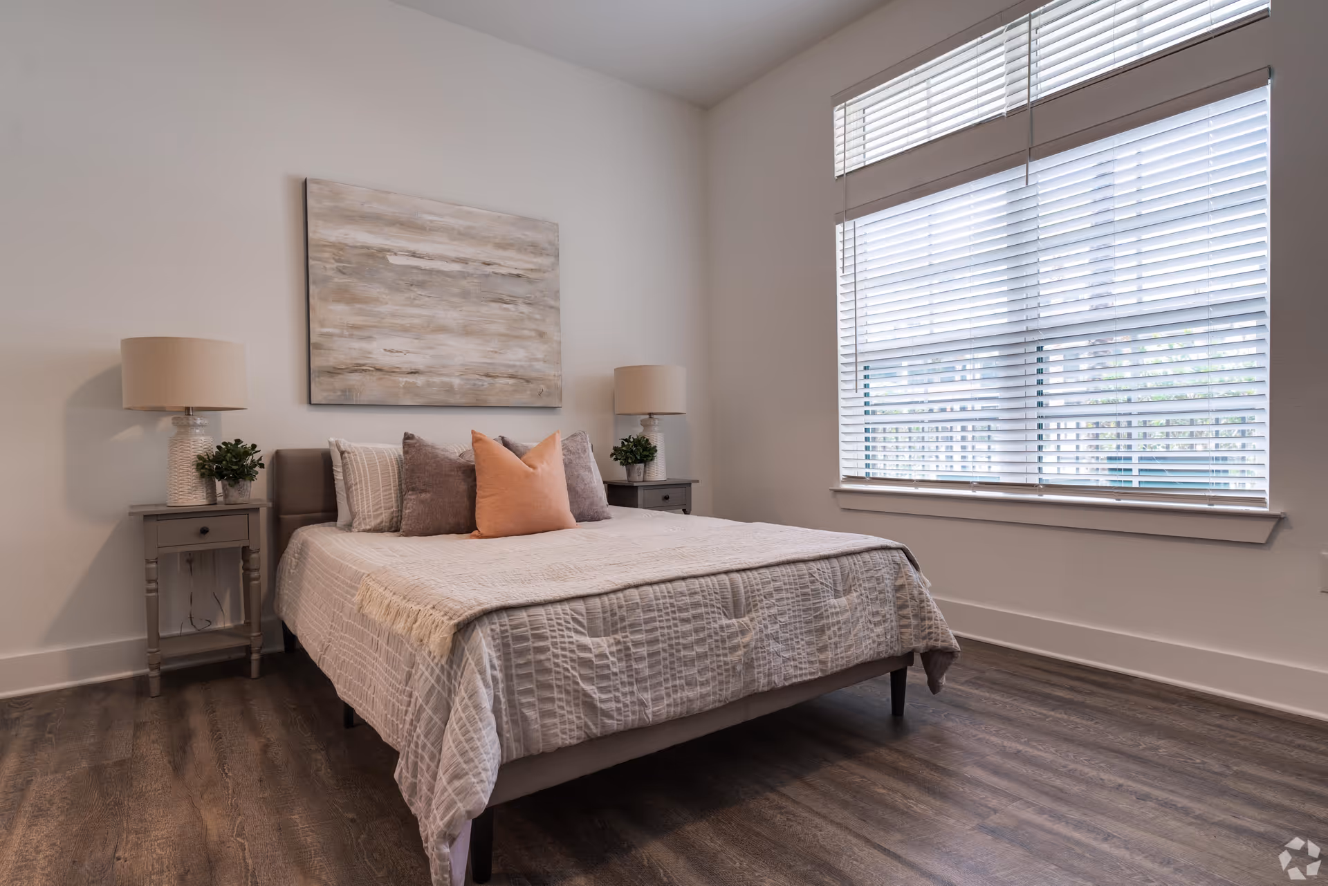 A neatly made bed with a beige quilt and multiple pillows in a bedroom. The bed is flanked by two gray nightstands, each with a lamp and a small plant. Above the bed hangs a large abstract painting in neutral tones. A large window with white blinds allows natural light into the room, which has wood flooring and white walls.