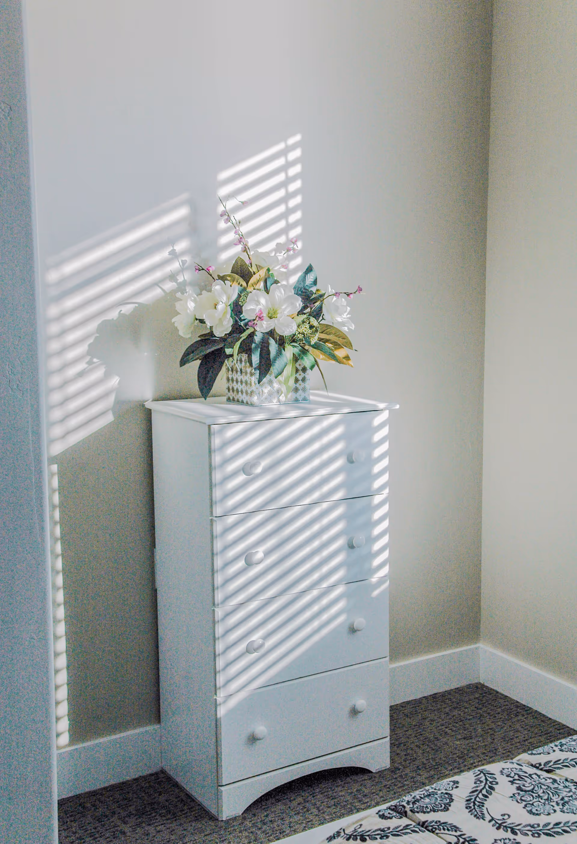 A white chest of drawers with four drawers stands against a beige wall. On top of the chest is a decorative vase with white and pink flowers and green leaves. Sunlight streams through window blinds, casting striped shadows on the chest and wall. Part of a bed with a patterned bedspread is visible in the lower right corner.