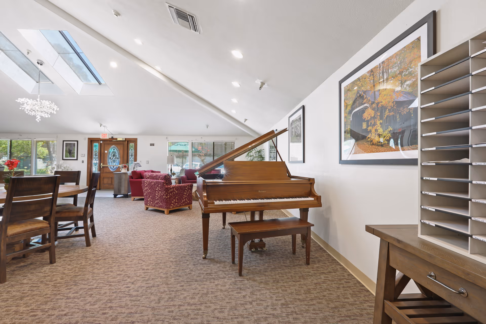 Interior view of a senior living facility common area featuring a grand piano with a bench, a wooden dining table with chairs, and comfortable seating with red upholstered armchairs. The room has a slanted ceiling with skylights, framed artwork on the walls, and large windows letting in natural light. There is also a wooden table with a mail organizer on the right side.