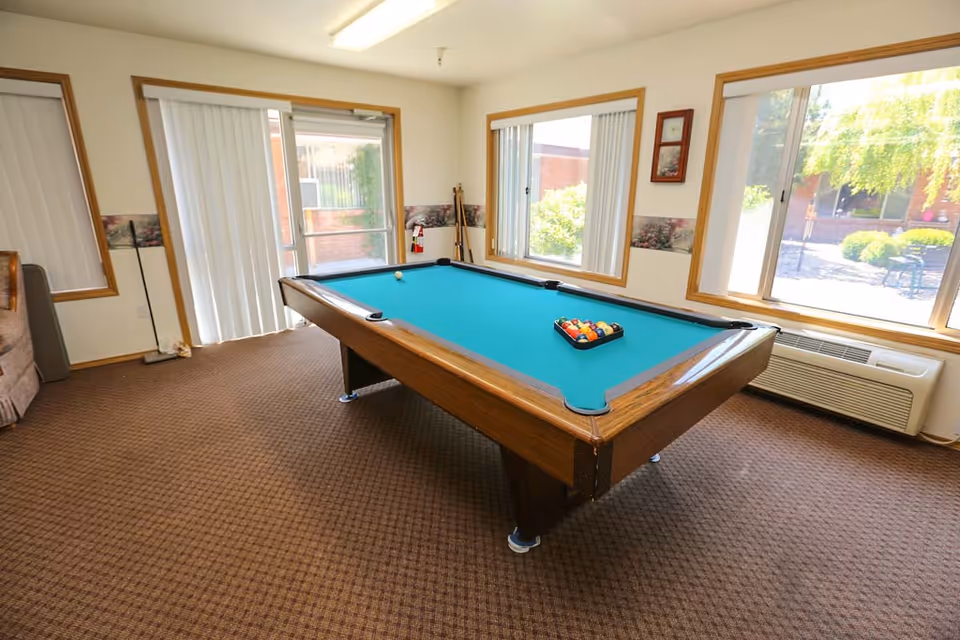 A room with a pool table featuring a blue felt surface, set up with billiard balls and a cue ball. The room has large windows with vertical blinds allowing natural light to enter, beige walls with a decorative border, and brown carpeted flooring. There is a fire extinguisher in the corner and an air conditioning unit beneath one window.