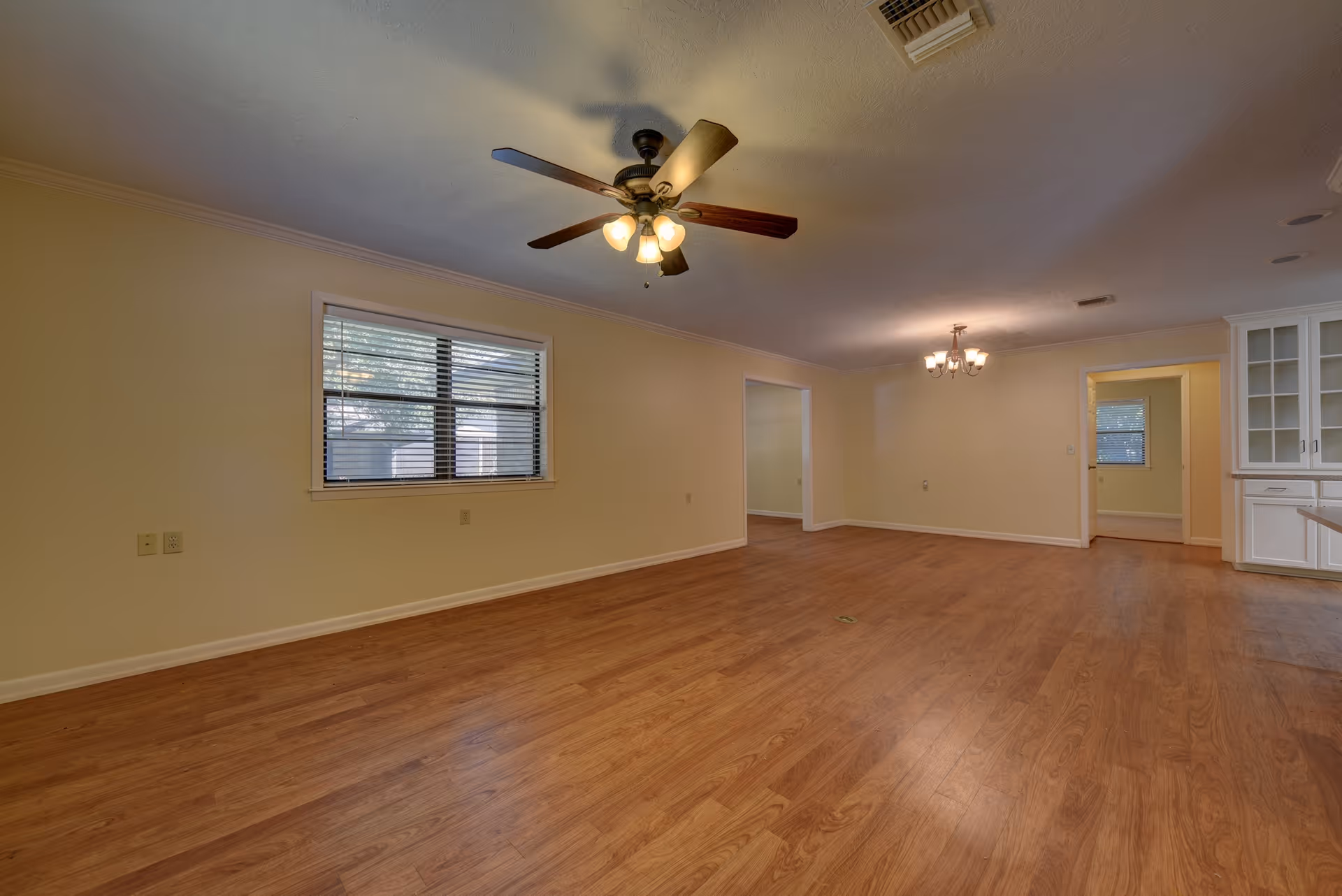 Empty room with wooden flooring, beige walls, a ceiling fan with lights, a window with blinds, and a chandelier light fixture. There is a doorway leading to another room and built-in white cabinetry on the right side.