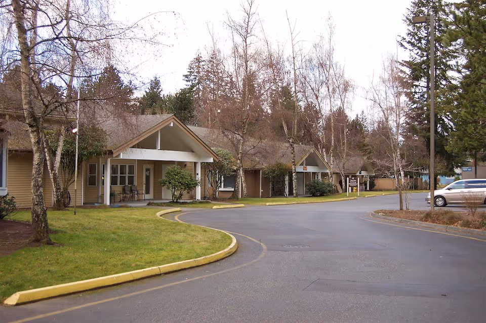 Exterior view of a senior living facility named Pacifica Dogwood Cottage showing a curved driveway, several single-story buildings with covered entrances, leafless trees, and a parked silver vehicle on the right side.