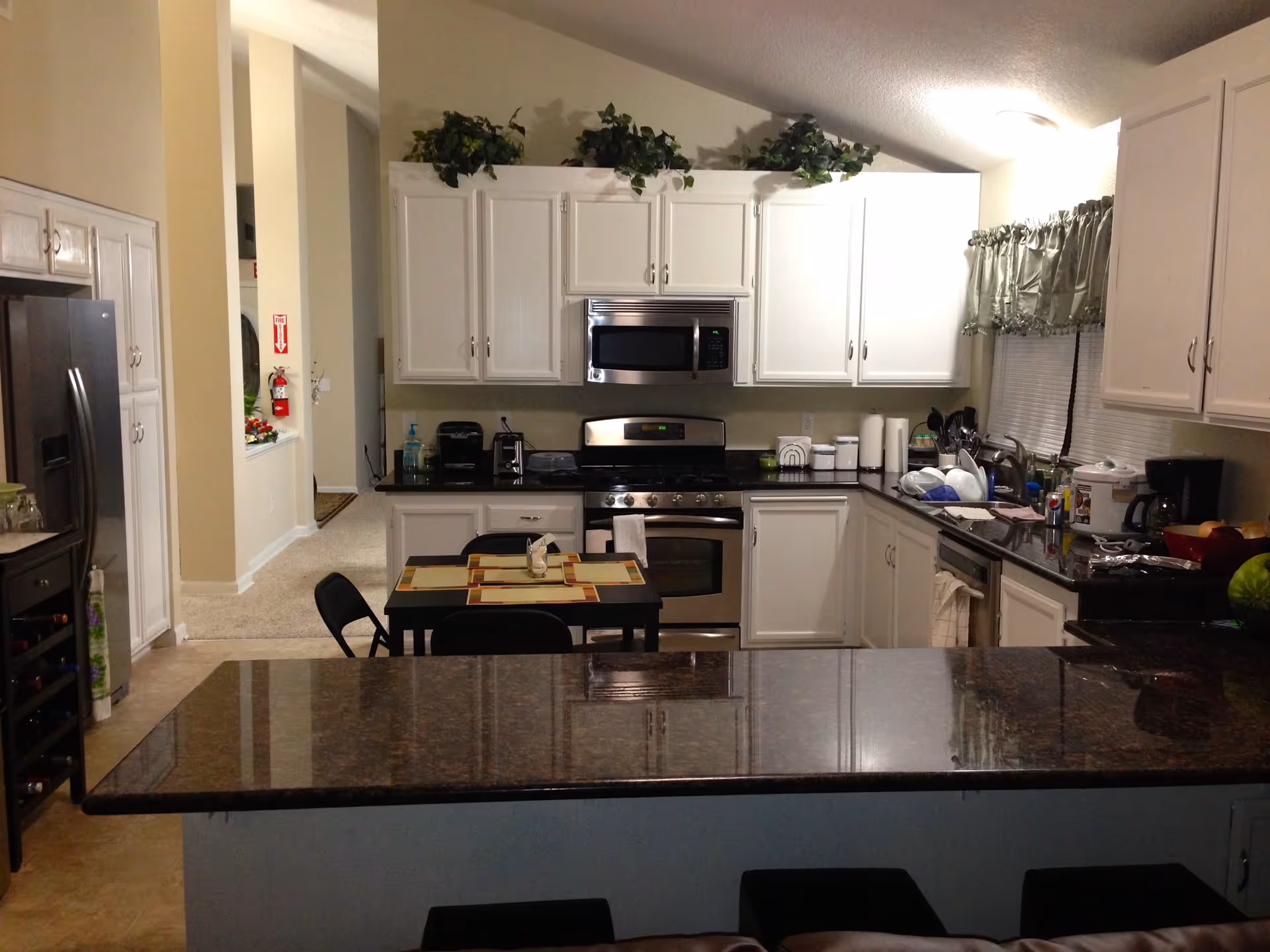 Interior view of a kitchen with white cabinets, a stainless steel microwave and stove, a black countertop with various kitchen appliances and utensils, a small dining table with four chairs, and a granite kitchen island in the foreground.