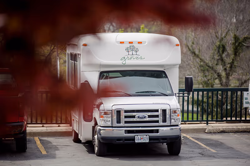 A white shuttle bus with the logo 'The Groves' on the front, parked in a parking lot with a red vehicle partially visible on the left. The background shows a green railing and trees with no leaves, indicating a late autumn or early spring setting.