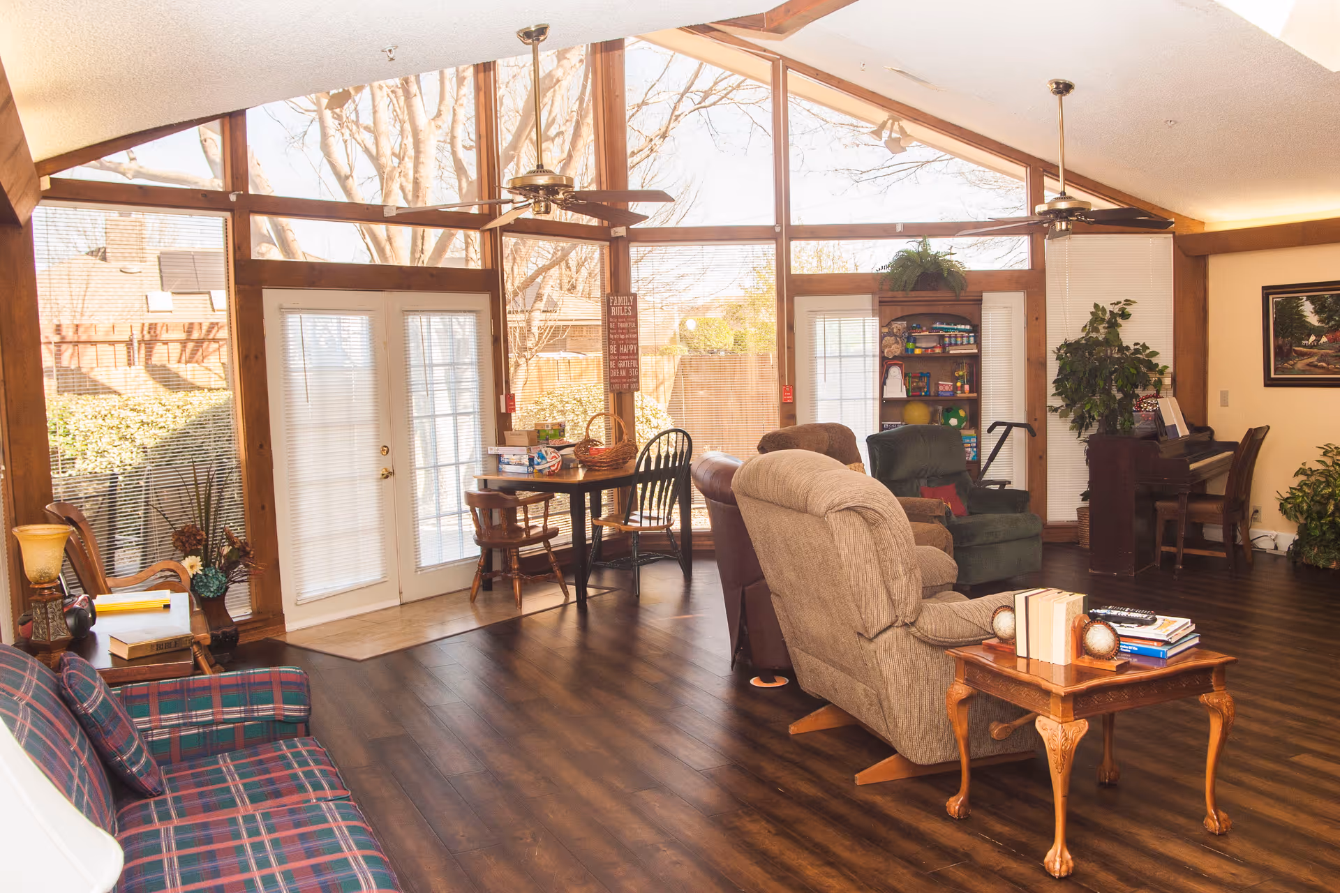 A bright and cozy living room with large floor-to-ceiling windows and glass doors letting in natural light. The room features a plaid sofa, two recliners, a small wooden table with books, a round dining table with chairs, a piano, and several plants. The ceiling has two ceiling fans and wooden beams, and the floor is dark wood.