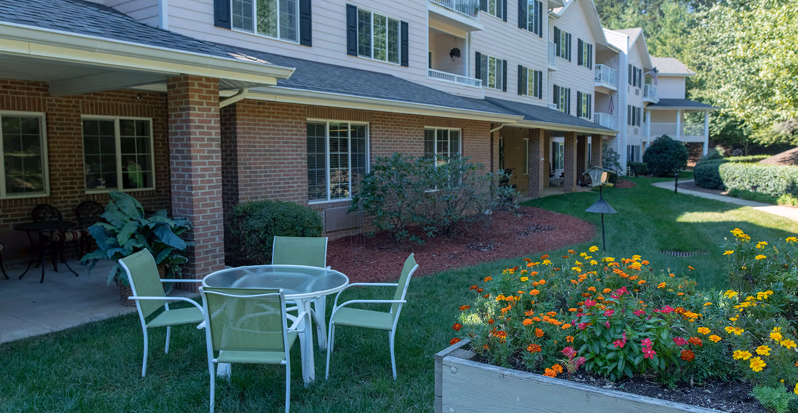 Outdoor seating area with a round glass table and four green chairs on a grassy lawn next to a flower bed with colorful flowers. The background shows a multi-story building with brick and white siding, windows with shutters, and a covered patio area with additional seating.