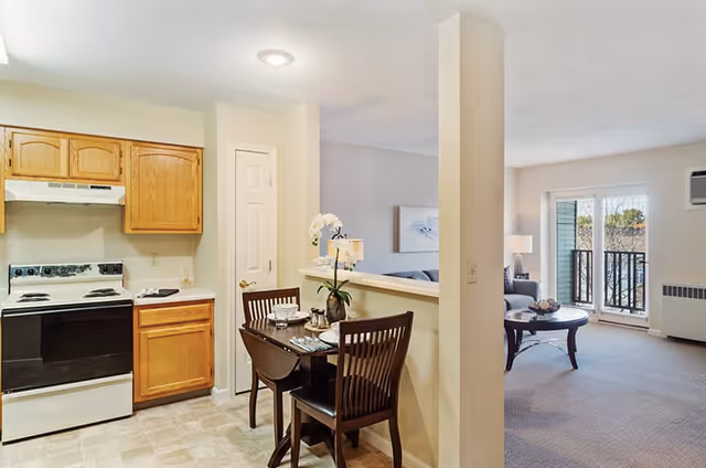 Open-plan interior showing a small kitchen with wooden cabinets, a two-seat dining table, and a living room with balcony doors.