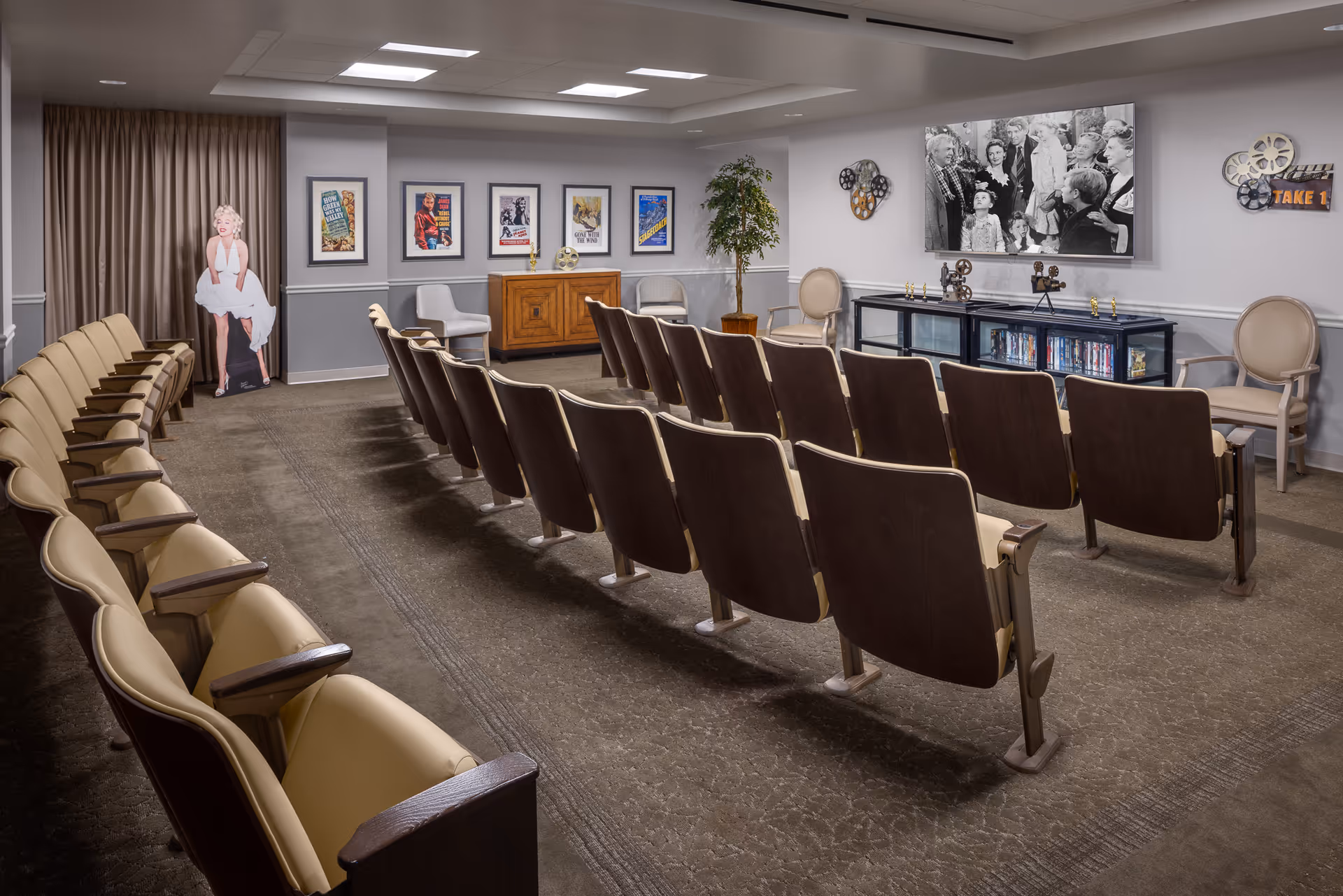 A small theater room with rows of beige and brown theater seats facing a wall-mounted black and white movie scene. The room has movie-themed decorations including framed vintage movie posters, film reels, and a cardboard cutout of Marilyn Monroe. There are also a few beige chairs and a cabinet with DVDs underneath the screen.