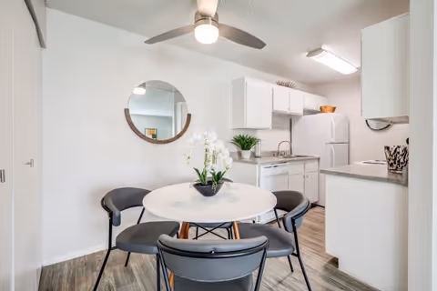 A bright and clean kitchen and dining area featuring white cabinets, a refrigerator, a sink, and a round white dining table with four black chairs. A decorative round mirror hangs on the wall above the table, and a potted white orchid is placed in the center of the table. The flooring is wood-style laminate.