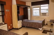 A nursing facility room with a single bed covered in a patterned blanket, a window with blinds and an air conditioning unit, a small table and chair, and a privacy curtain partially drawn around the bed area.