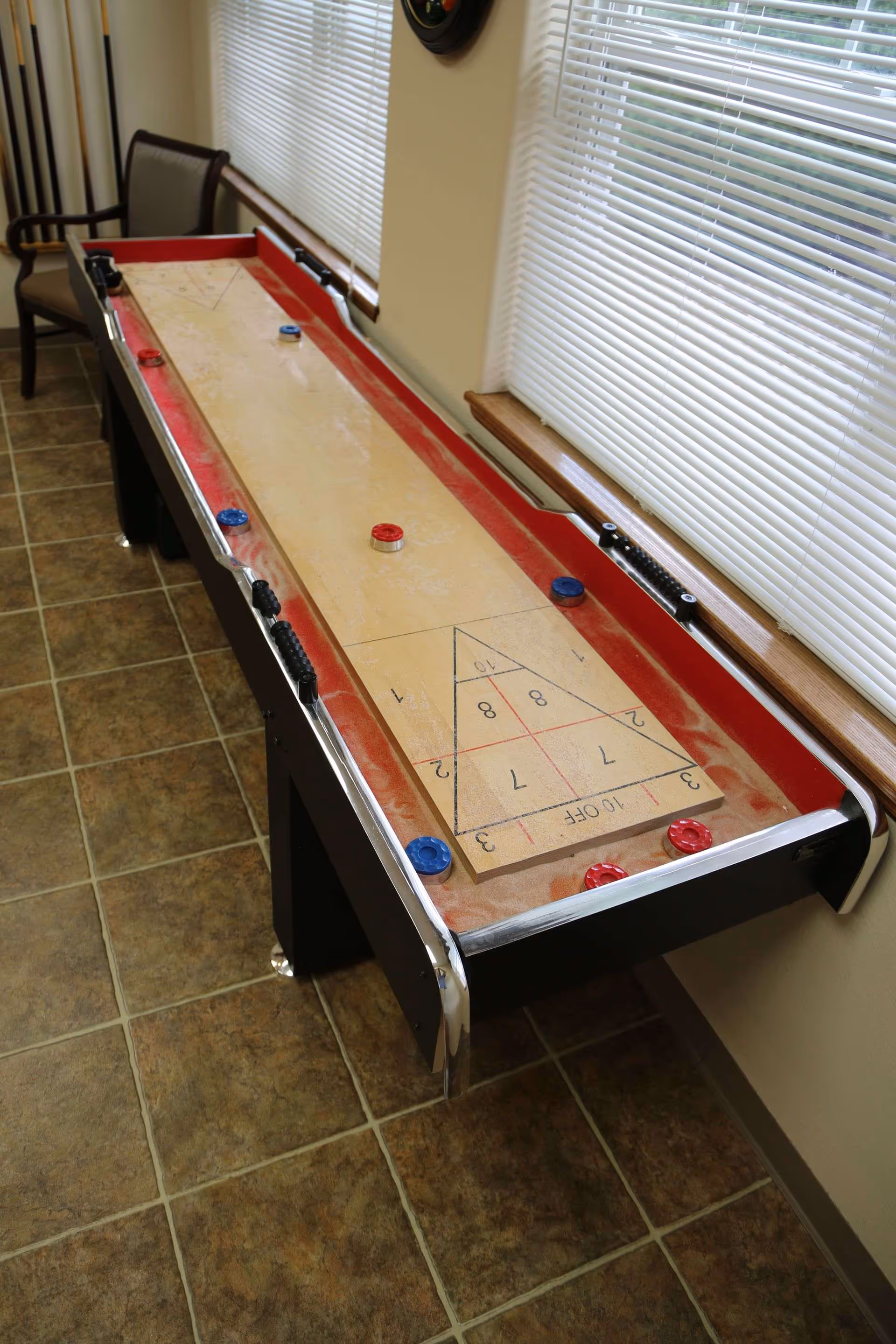 A shuffleboard table with red and blue pucks sits next to blinds-covered windows in a tiled room.