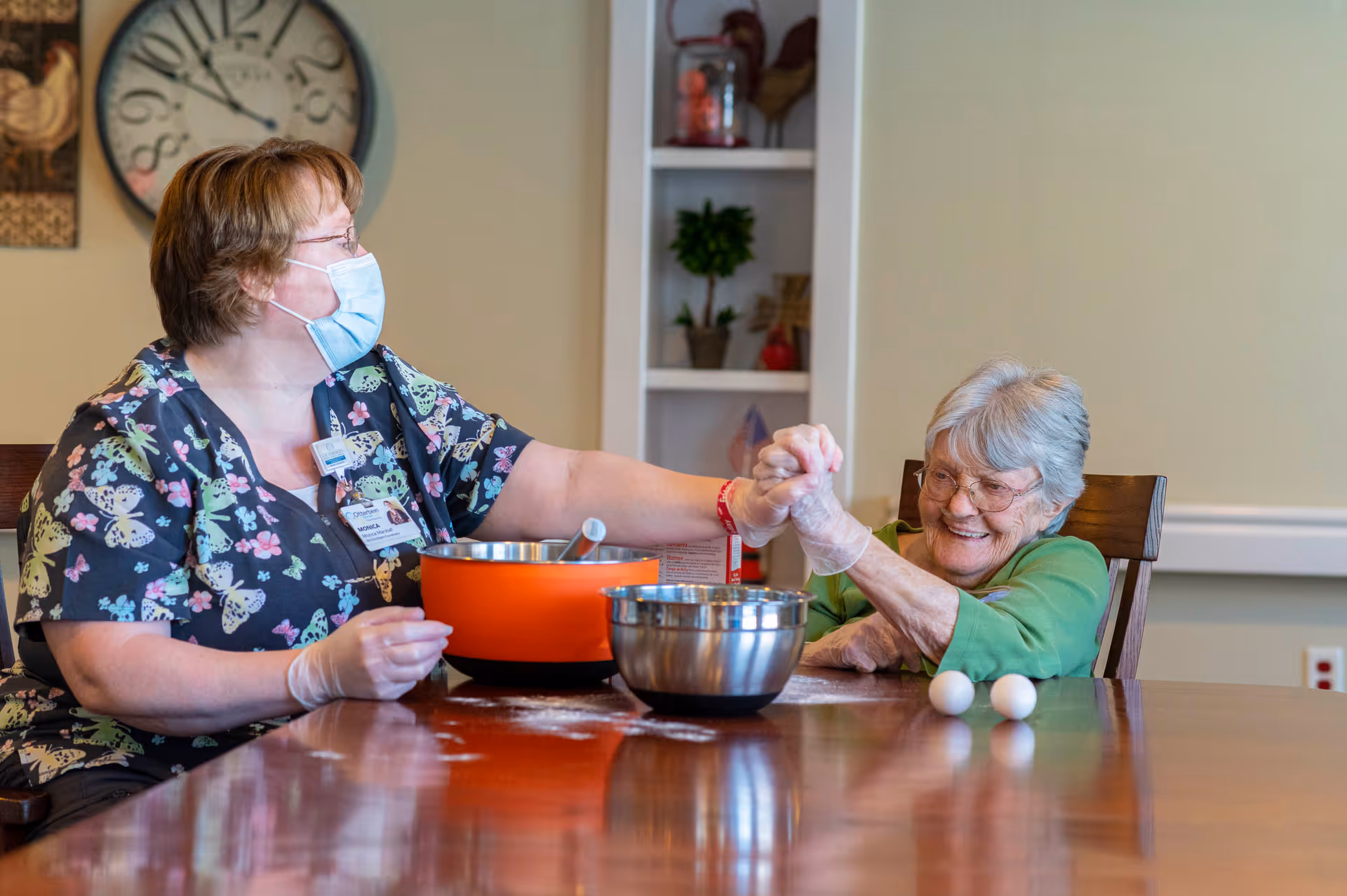 A caregiver wearing a mask and gloves is sitting at a table with an elderly woman. They are holding hands and smiling. On the table are two mixing bowls and two eggs, suggesting a cooking or baking activity in progress.