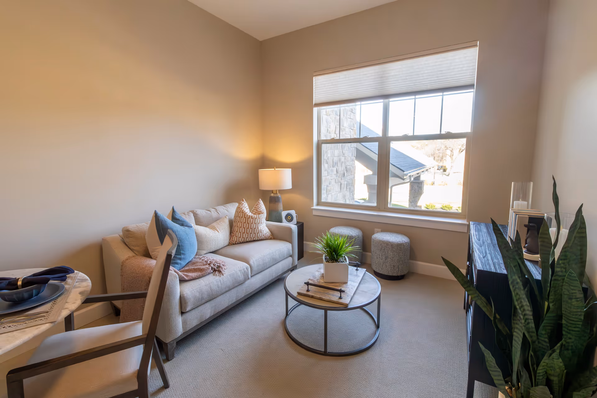 A cozy living room with a beige sofa adorned with multiple pillows, a round coffee table with a small plant, two gray ottomans near a large window with a view outside, a lamp on a side table, a dining chair partially visible, and a dark wooden cabinet with decorative items and a tall green plant beside it.