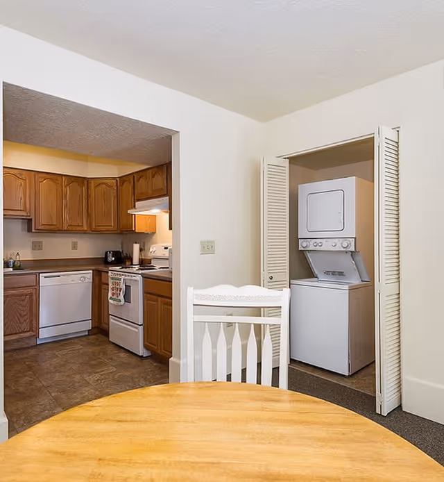 View of a kitchen area with wooden cabinets, a white stove, dishwasher, and a countertop. In the foreground, there is a round wooden table with a white chair. To the right, there is a small laundry closet with a stacked washer and dryer behind bi-fold doors.