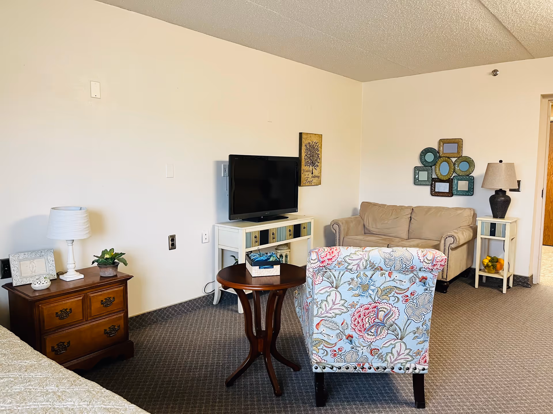 A cozy living room area in a senior living facility with a beige couch, a floral patterned armchair, a small round wooden table, a TV on a white stand, two side tables with lamps, and decorative wall art.