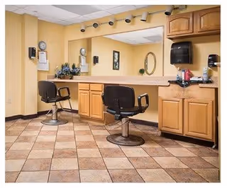 Interior view of a salon area in a senior living facility with two black salon chairs in front of a large mirror, wooden cabinets, a paper towel dispenser, and various hair care items on the counter. The floor is tiled with a brown and beige pattern.
