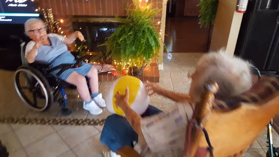 Two elderly women playing with a beach ball indoors. One woman is seated in a wheelchair wearing a light blue dress and white shoes, while the other woman is sitting in a wooden rocking chair. There is a fireplace with a green hanging plant and string lights in the background.