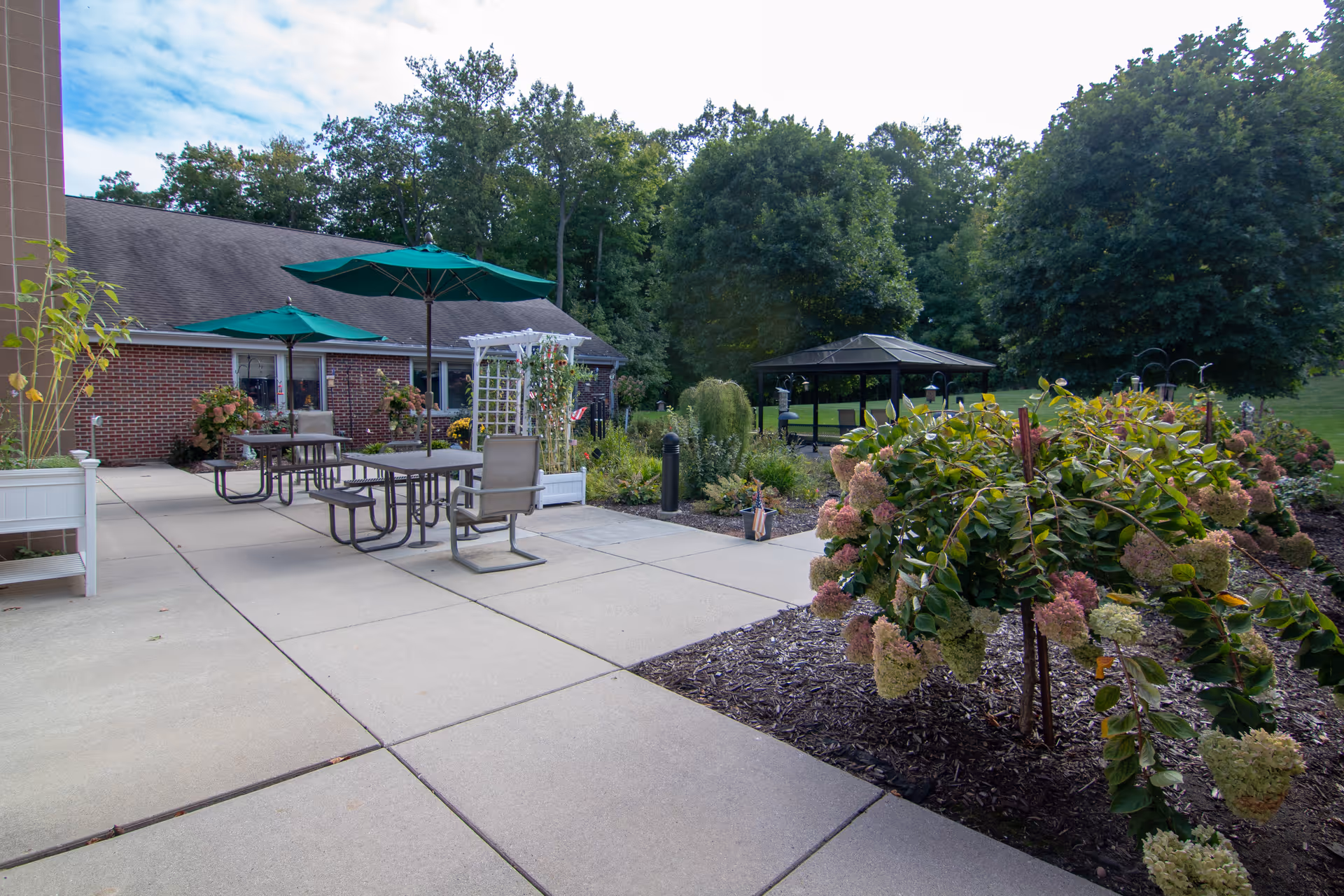Outdoor patio area at Rocky Knoll Health Care Center featuring tables with green umbrellas, chairs, flower beds with blooming plants, and a gazebo in the background surrounded by trees and greenery.