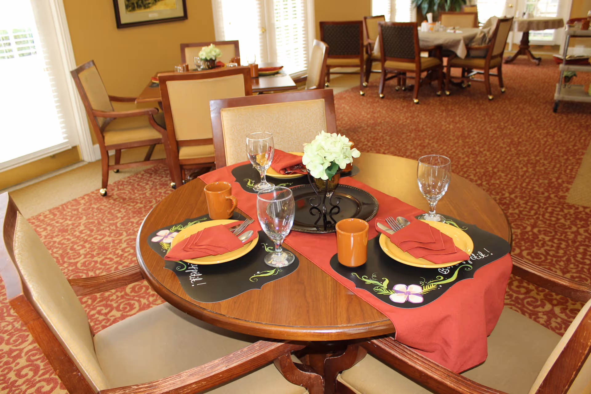 A round wooden dining table set for two with yellow plates, red napkins, brown mugs, and clear water glasses. A small flower arrangement in a black vase is in the center on a red table runner. The room has patterned red carpet and several other tables and chairs in the background near large windows with white blinds.