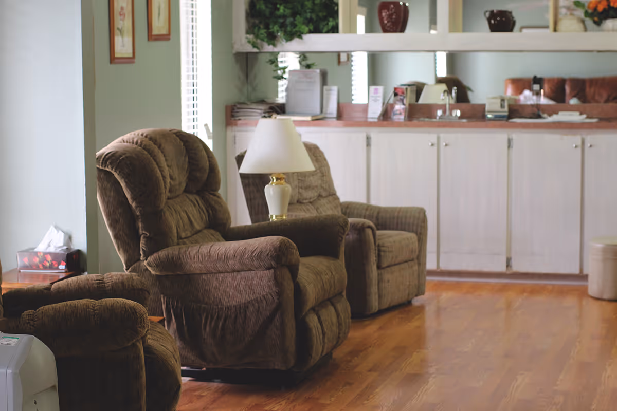 A cozy living room area with three brown upholstered recliner chairs arranged near a white cabinet with a countertop. A white table lamp is placed on a small table between two of the chairs. The room has wooden flooring and some framed pictures on the wall.