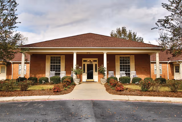 Single-story brick building with a covered columned front porch, rocking chairs, landscaped shrubs, and a driveway.
