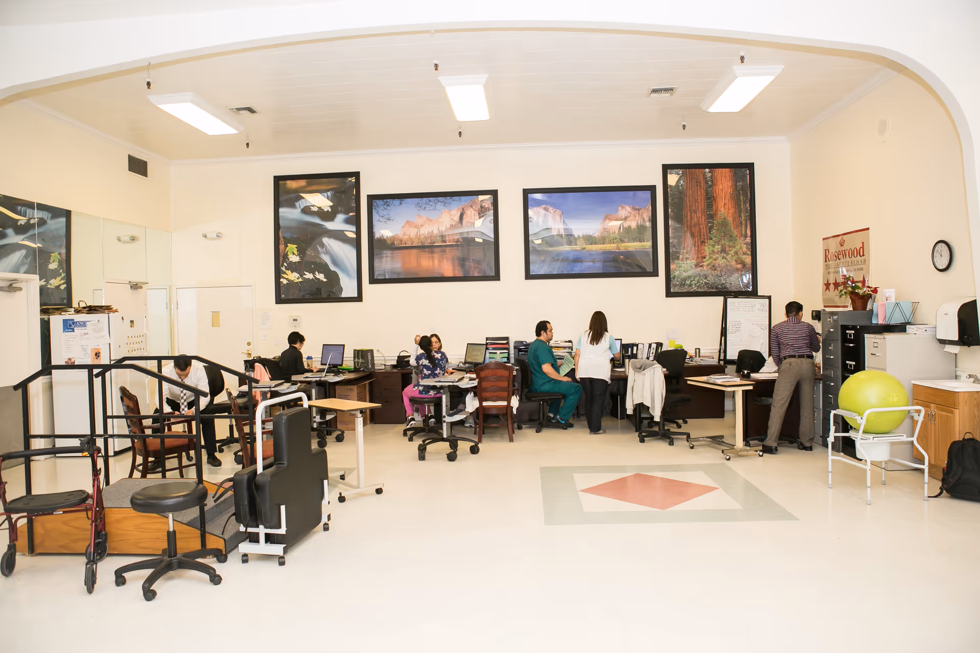 Open interior workspace at a senior facility with desks, medical/therapy equipment and staff working beneath large landscape photos.