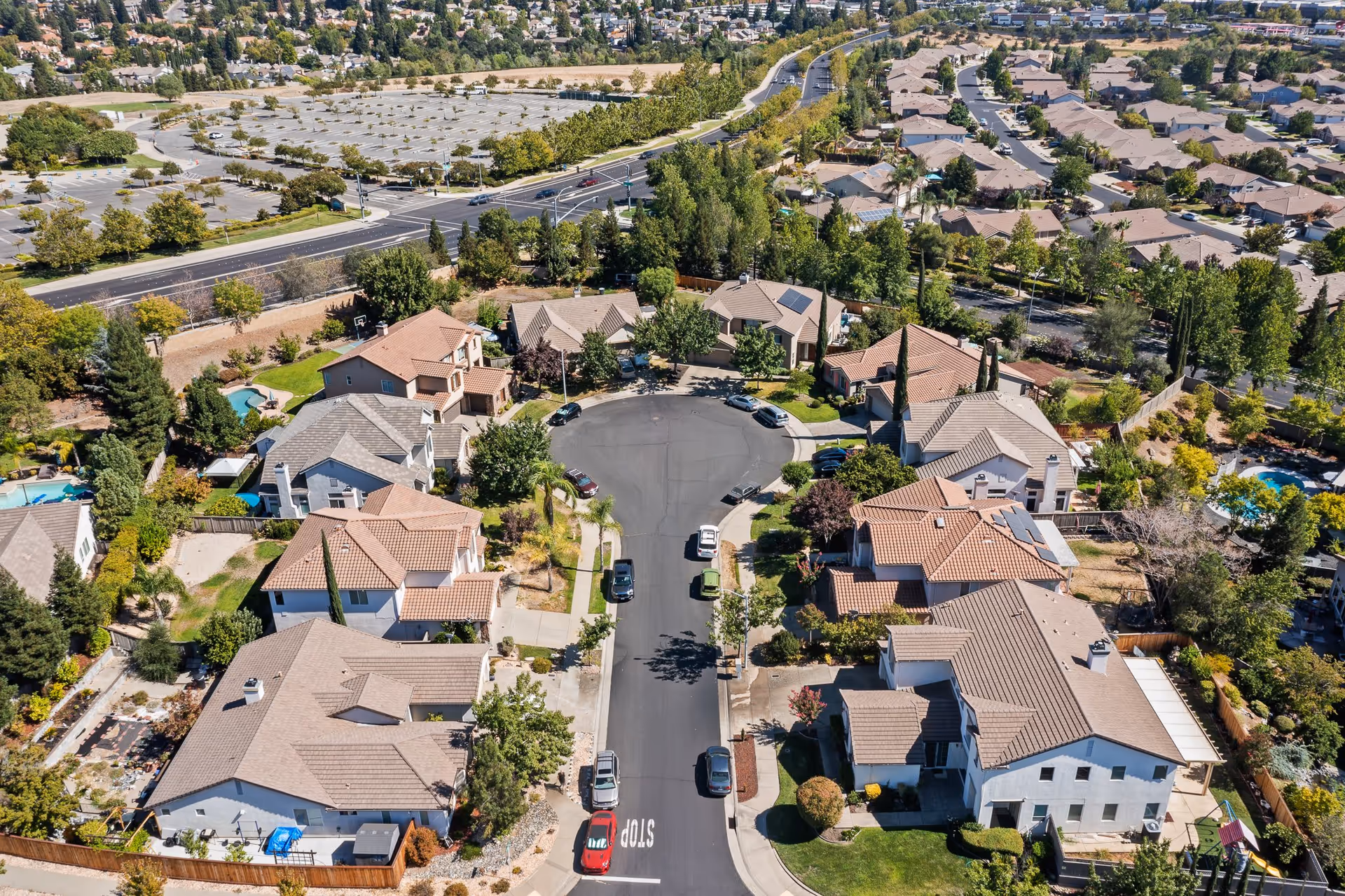 Aerial view of a suburban residential cul-de-sac with multiple houses, parked cars, trees, and well-maintained yards. Surrounding the neighborhood are more houses, roads, and a large empty parking lot in the background.